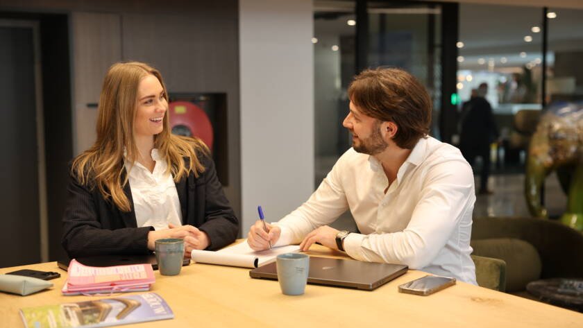 Man en vrouw zittend aan tafel, man schrijft terwijl vrouw lacht
