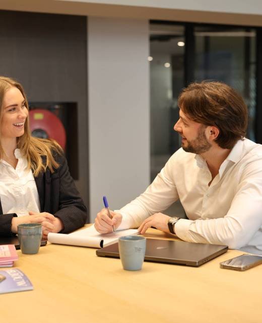 Man en vrouw zittend aan tafel, man schrijft terwijl vrouw lacht
